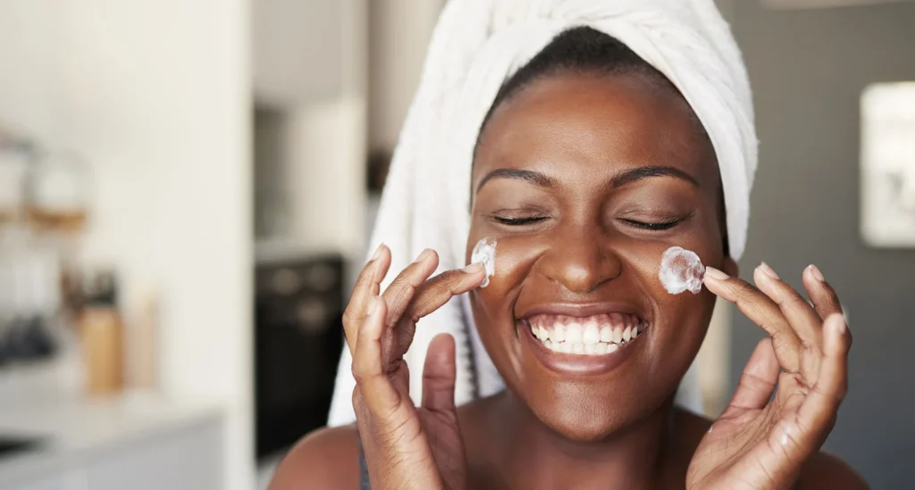 Black Woman smiling as she is putting on skincare or moisturizer 